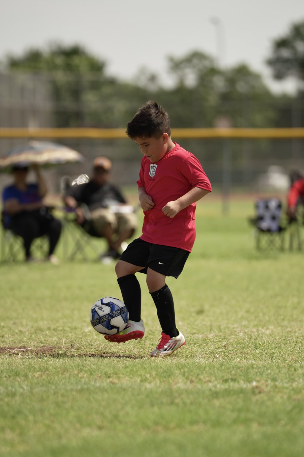 Young player in red taking a touch on the ball, parents watching from the sideline